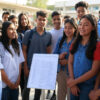 A person in a suit holds a sign reading "CBSE Results 2025" in front of a group of students in school uniforms, standing in an outdoor school courtyard decorated with balloons.